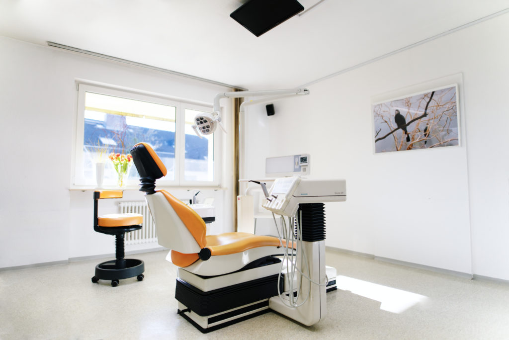 Ein Behandlungszimmer mit orangener Liege, Bild an der Wand und großem, sonnigen Fenster.